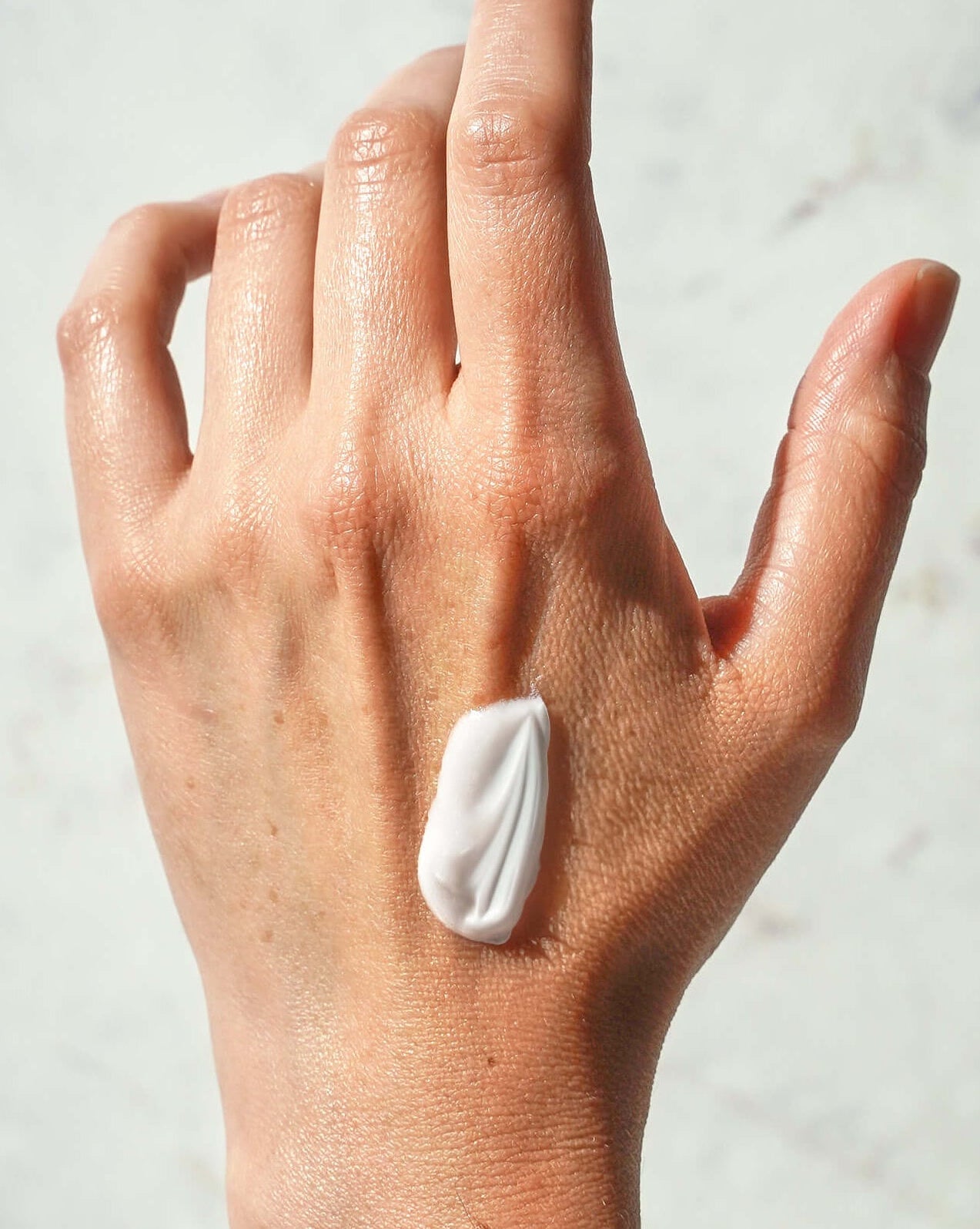 A close-up of a person's hand with a dollop of white cream or lotion applied to the back of the hand, set against a light gray marble-textured background.