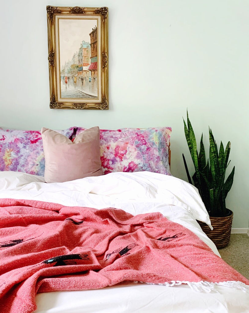 bedroom with multicoloured pillows and a coral thunderbird blanket draped over the end of the bed. A snake plant in a woven basket sits in the corner.